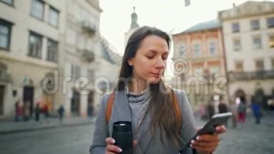 女人手里拿着一个<strong>保温</strong>杯，在夕阳下用智能手机沿着一条老街走着。 通信、社交网络